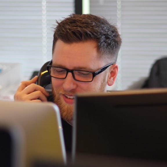 Man with beared and glasses talking on phone while smiling behind two computers.